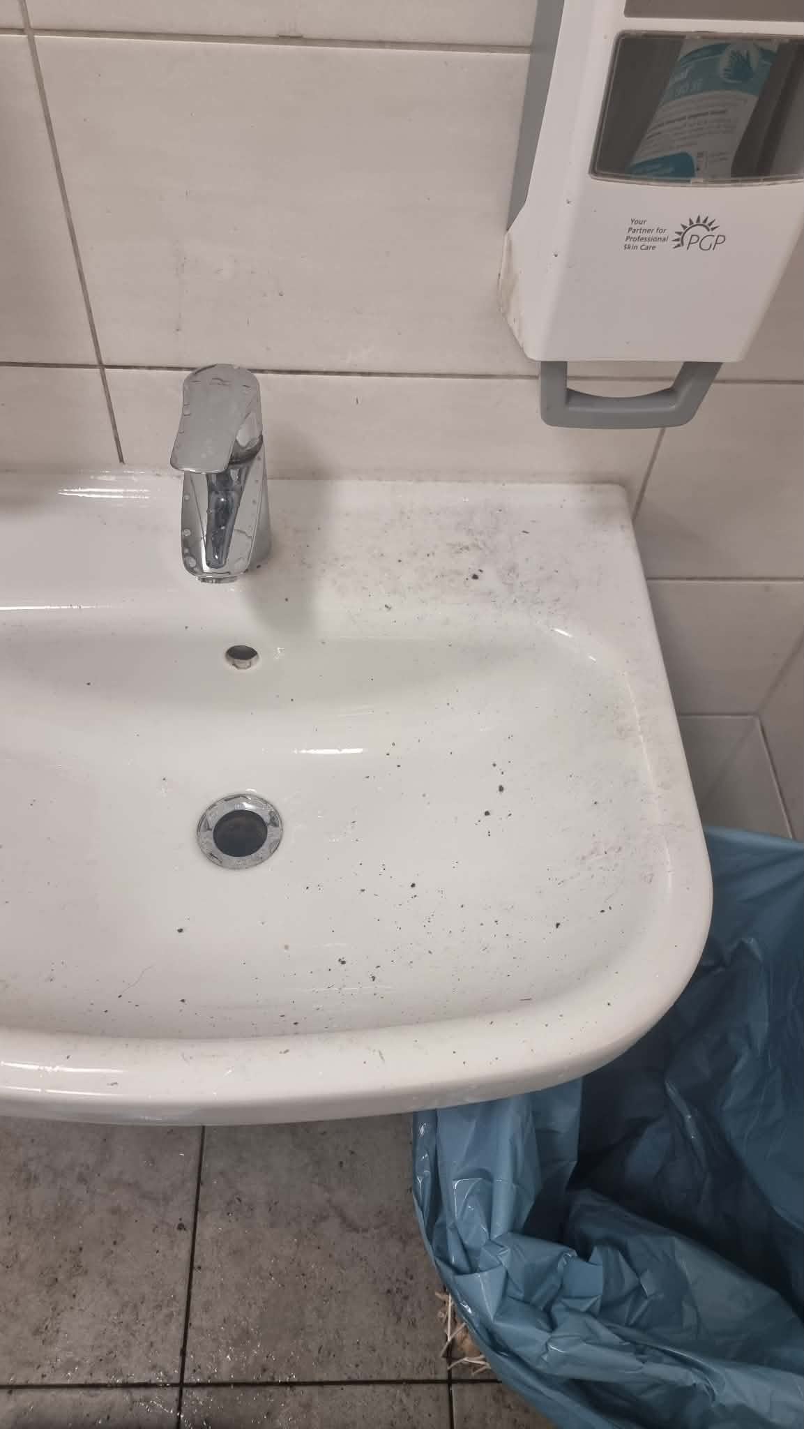 Waschbecken in einer Sanitäranlage vor der Reinigung, mit Ablagerungen im Becken und auf der Ablage